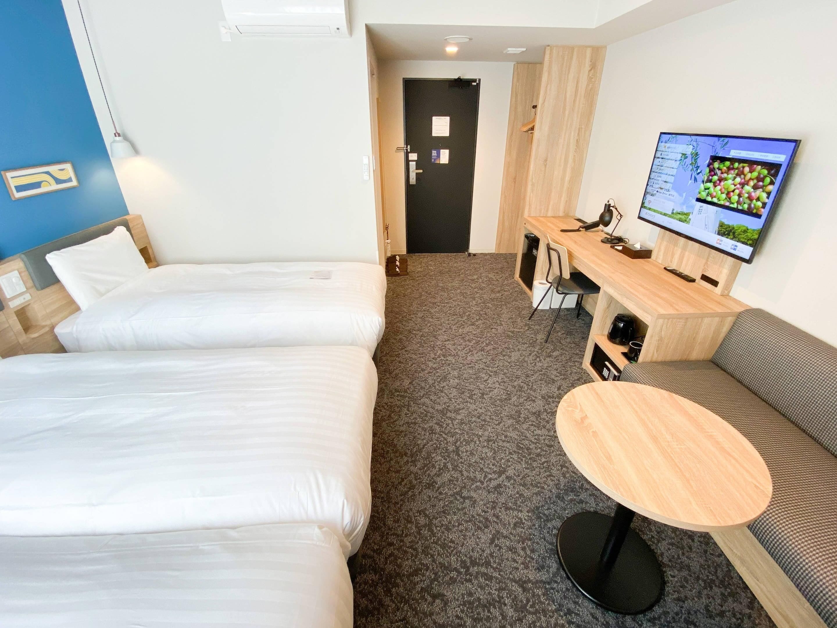 A hotel room with two beds, a desk with a computer and chair, a round table, and a TV on the wall.