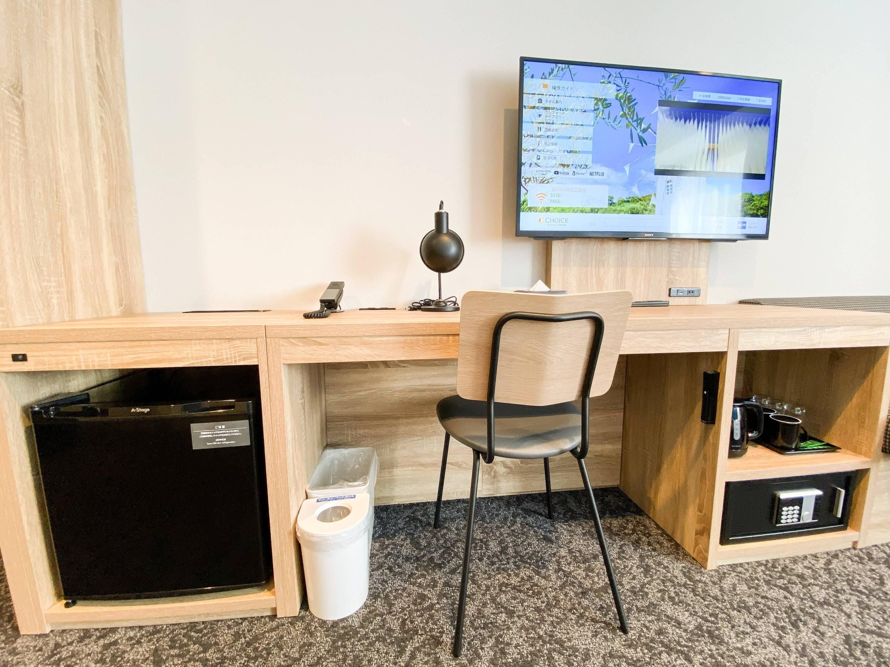 Quarto de hotel com escrivaninha de madeira, cadeira, televisão na parede, lixeira e mini geladeira.