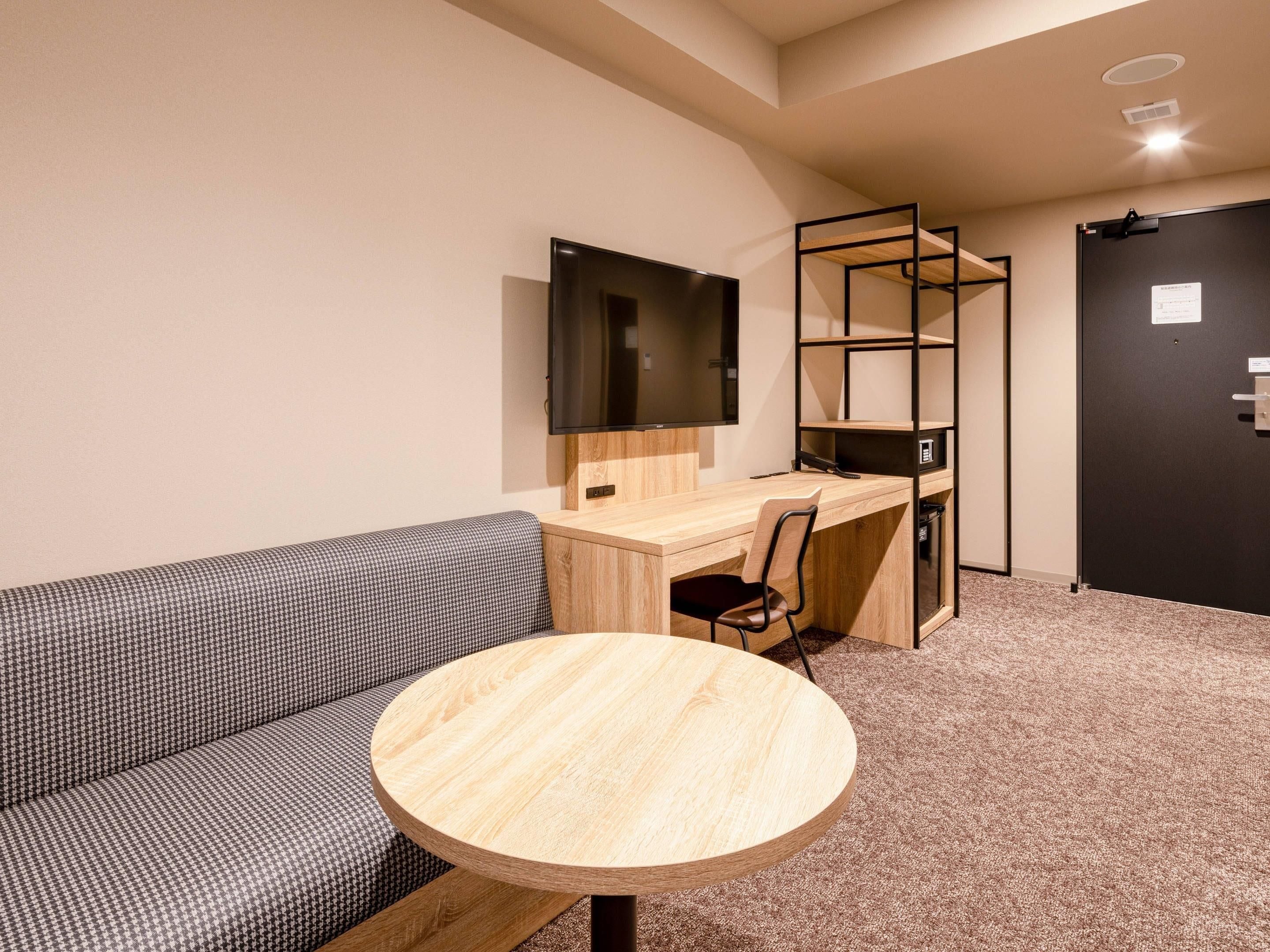 A room with a sofa, a desk with a chair, a round table, a TV mounted on the wall, and a door.