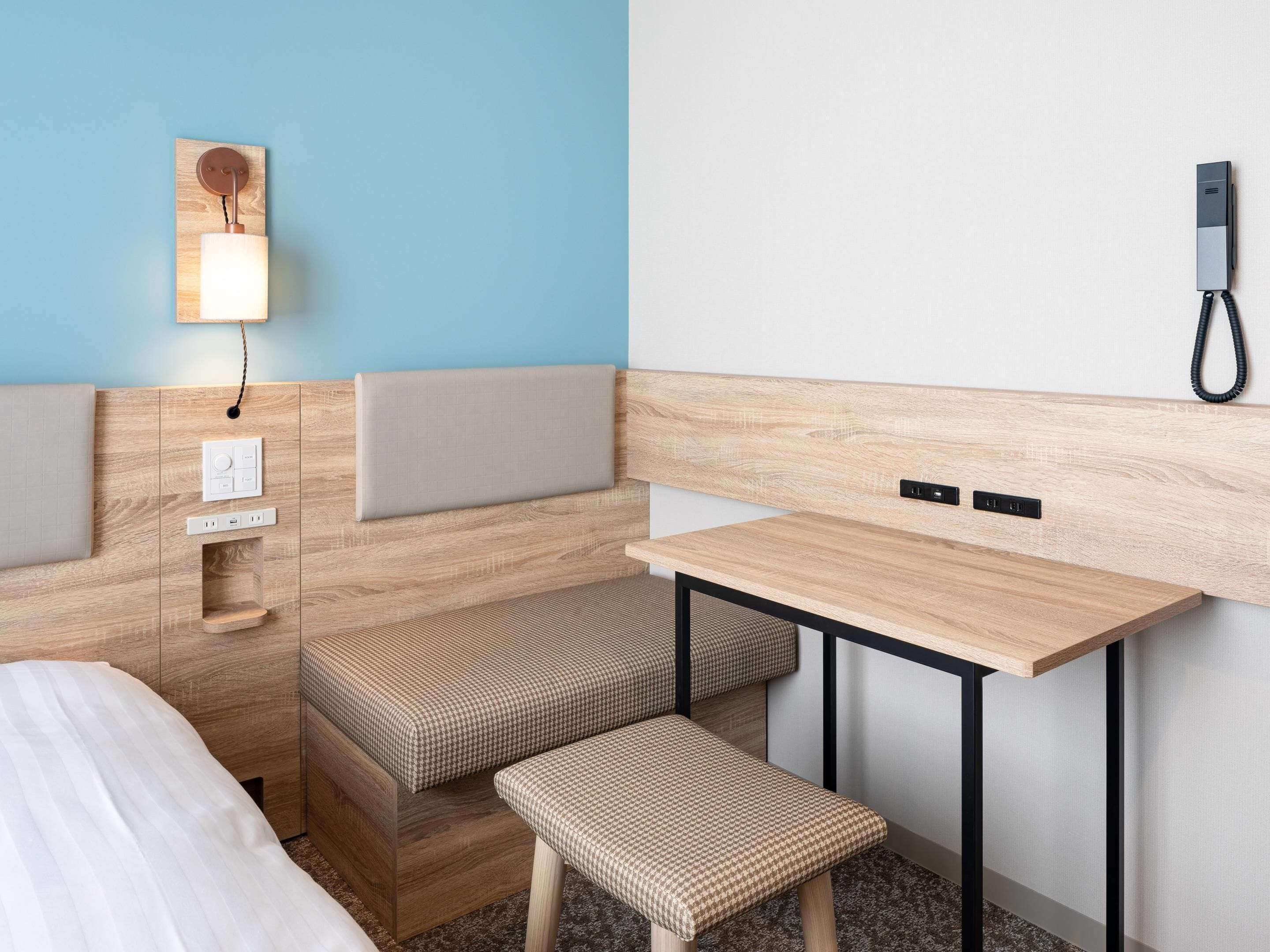A compact hotel room with a bed, a small desk, a bench, and a wall-mounted phone.