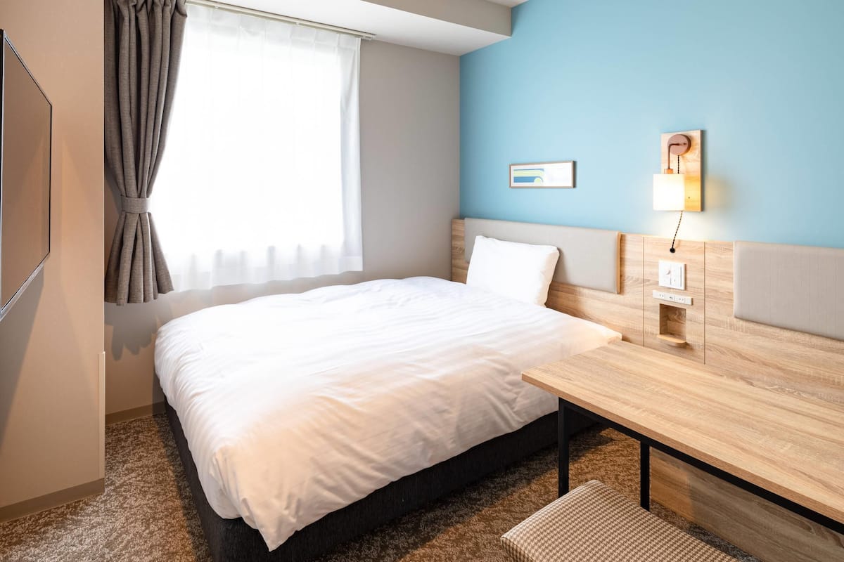 Una habitación de hotel con cama, escritorio, lámpara de pared y ventana con cortinas.
