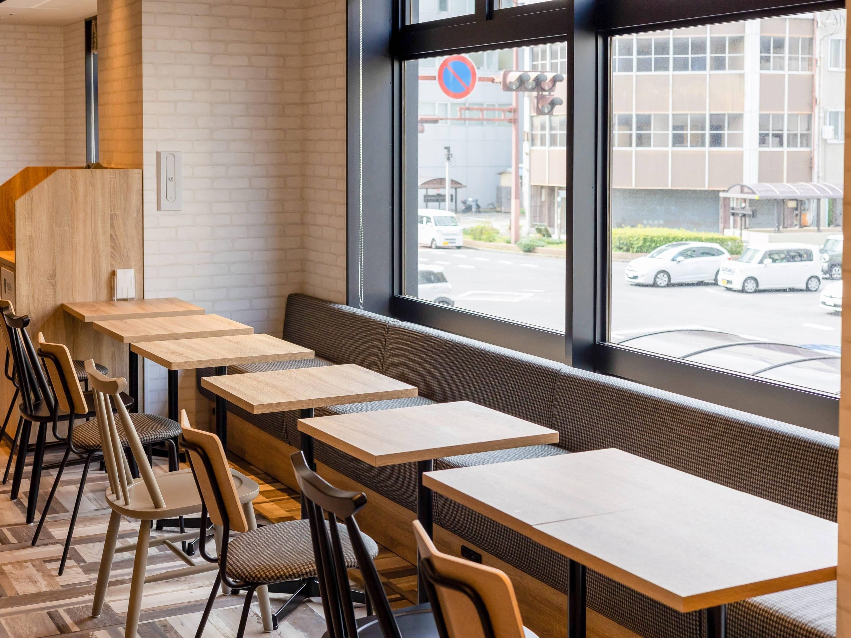 En modern caféinredning med träbord och stolar, en tegelvägg och utsikt över gatan genom stora fönster.