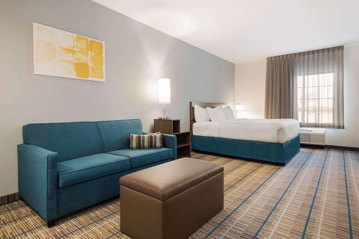 A hotel room with a blue sofa, a brown ottoman, a bed with white bedding, a nightstand, a lamp, and a window with curtains.