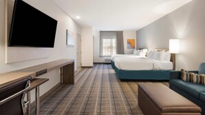 A hotel room with a flat-screen TV mounted on the wall, a bed with white bedding, a blue sofa, a bench, and a desk with a chair. - MainStay Suites Gaylord (Gaylord)