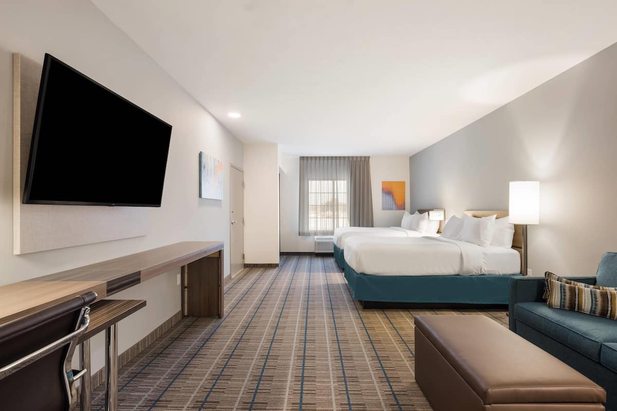 A hotel room with a flat-screen TV mounted on the wall, a bed with white bedding, a blue sofa, a bench, and a desk with a chair.