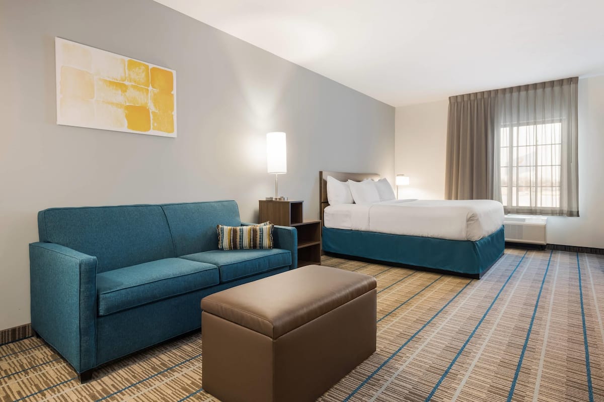 A hotel room with a blue sofa, a brown ottoman, a bed with white bedding, a nightstand, a lamp, and a window with curtains.