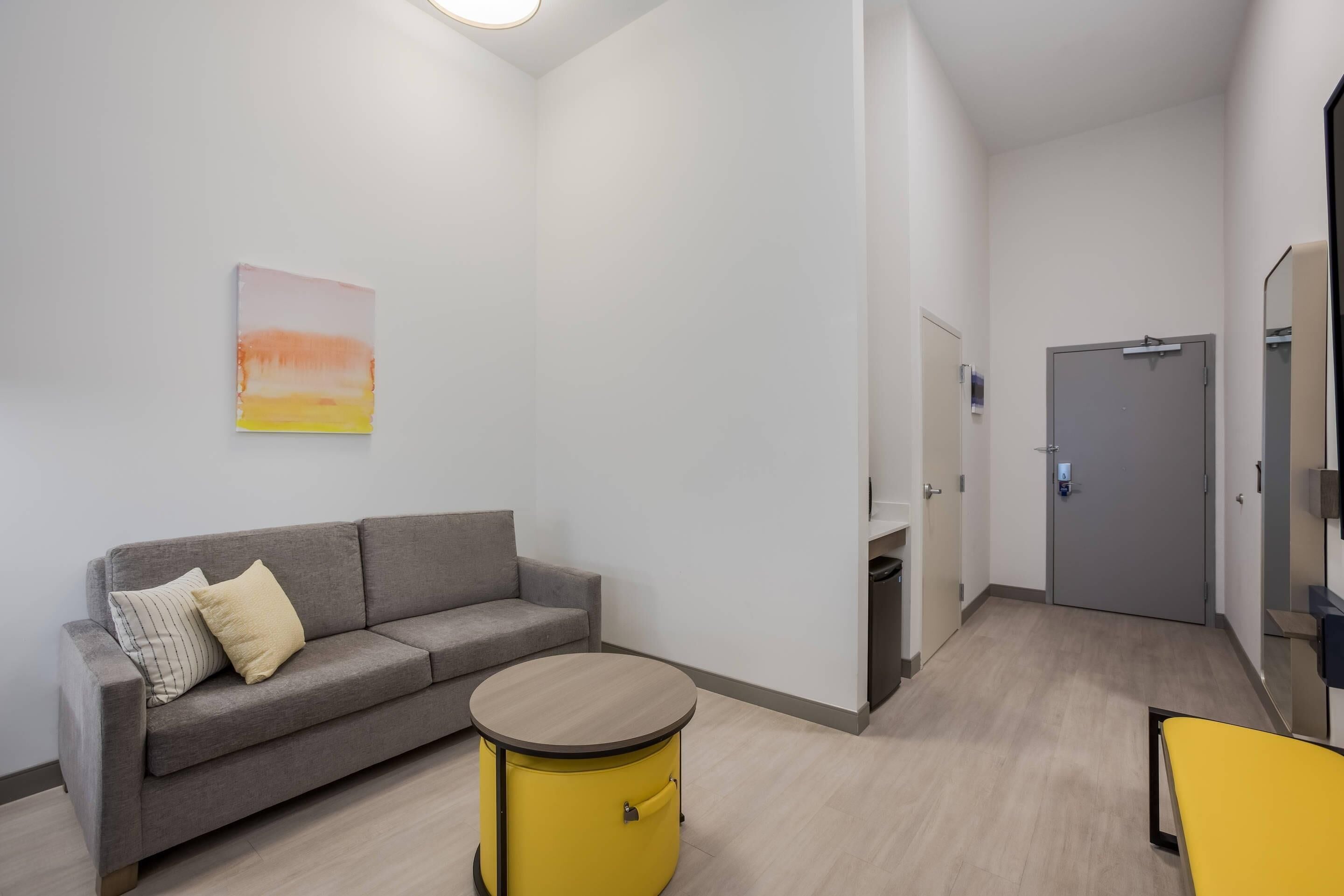 Una habitación moderna y minimalista con un sofá gris, un taburete amarillo tipo tambor y una puerta gris.