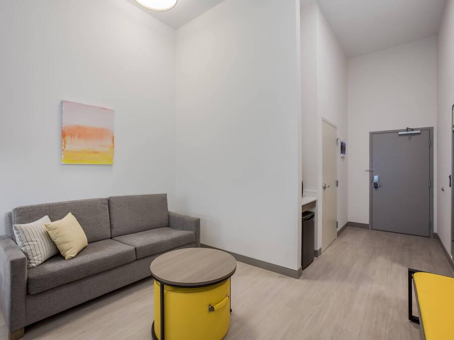 Una habitación moderna y minimalista con un sofá gris, un taburete amarillo tipo tambor y una puerta gris.