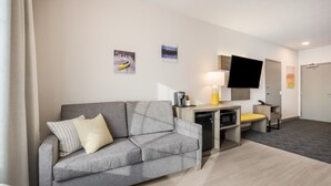 Une chambre d’hôtel moderne avec un canapé gris, un téléviseur à écran plat, un bureau avec une lampe et un four à micro-ondes.