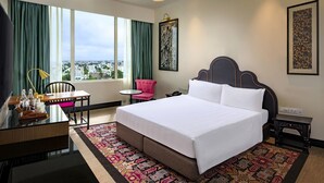 Habitación de hotel con una cama grande, un escritorio con silla, una mesita y vistas a la ciudad a través de amplios ventanales.