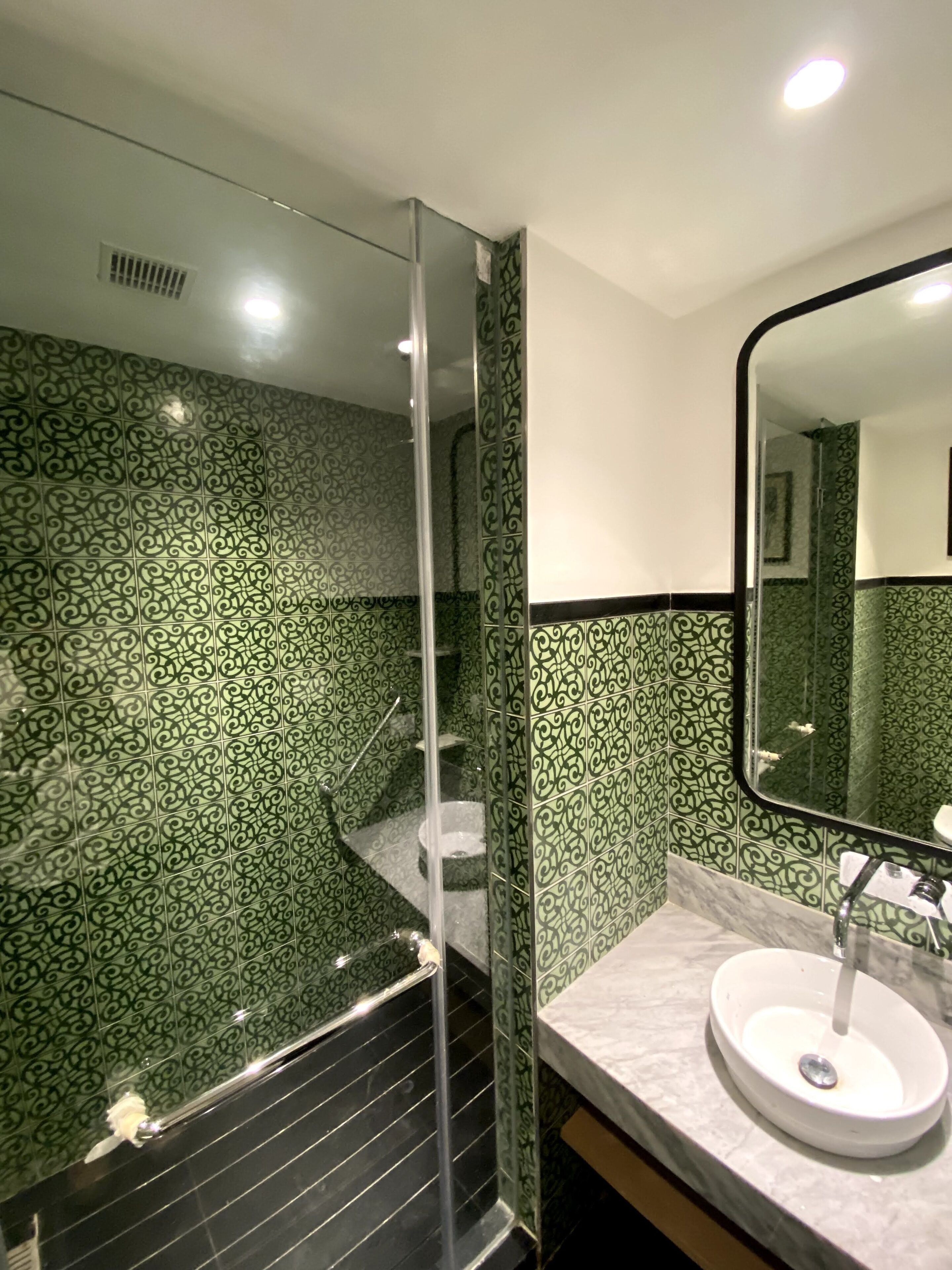 Een badkamer met groene betegeling, een douchecabine van glas, een witte wastafel en een spiegel.