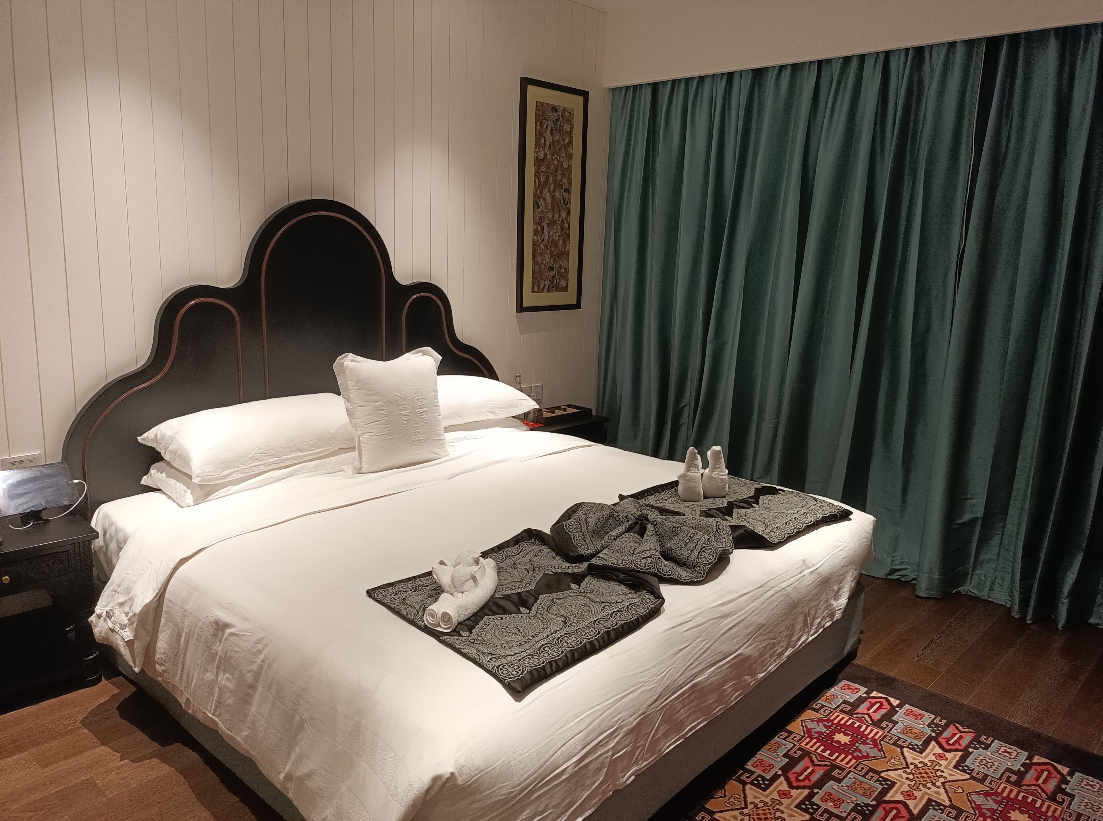 Cama bem arrumada com lençóis brancos, um manta decorativa e duas toalhas sobre um corredor estampado.