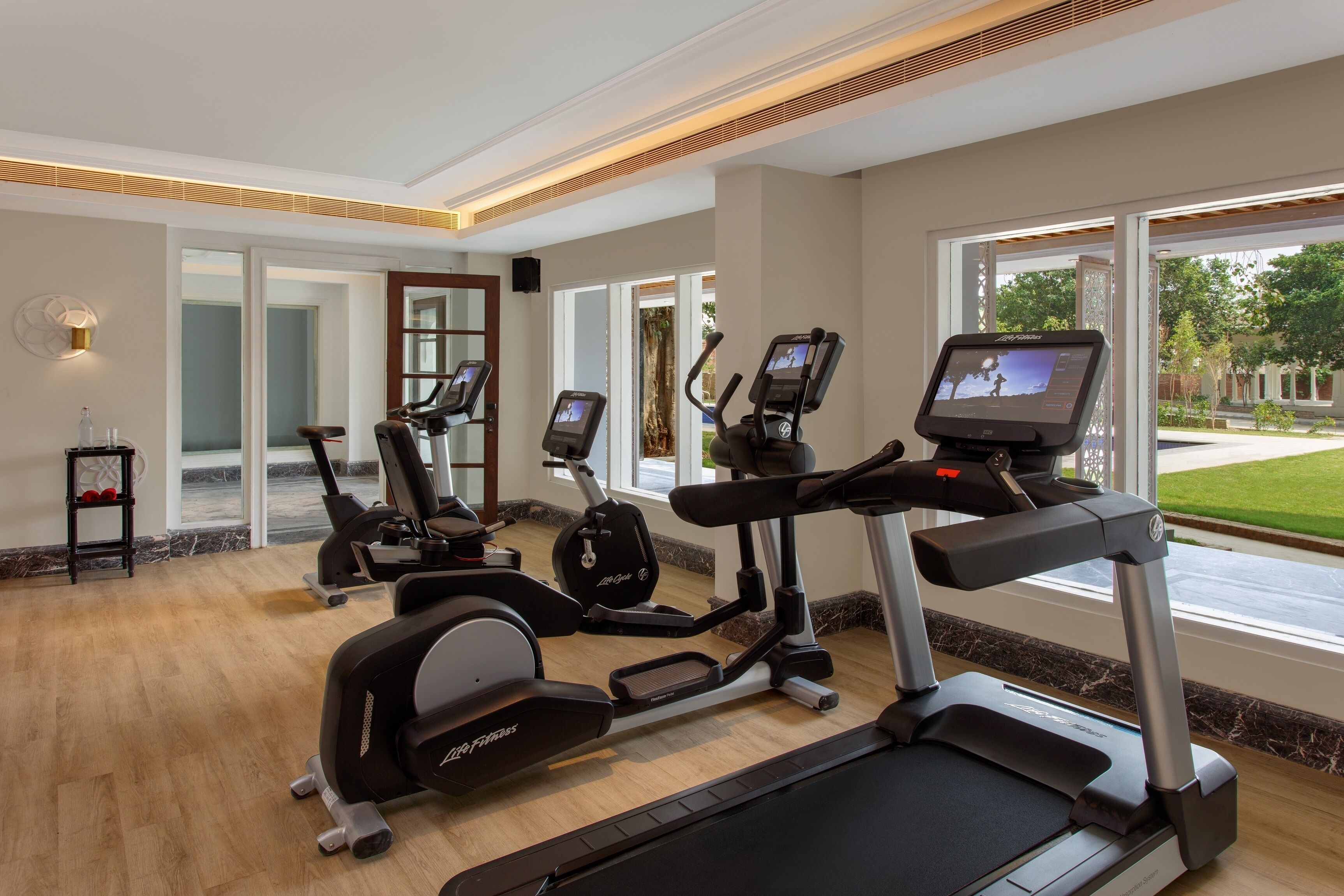 A gym with treadmills, ellipticals, and a stationary bike, large windows, and a wooden floor.