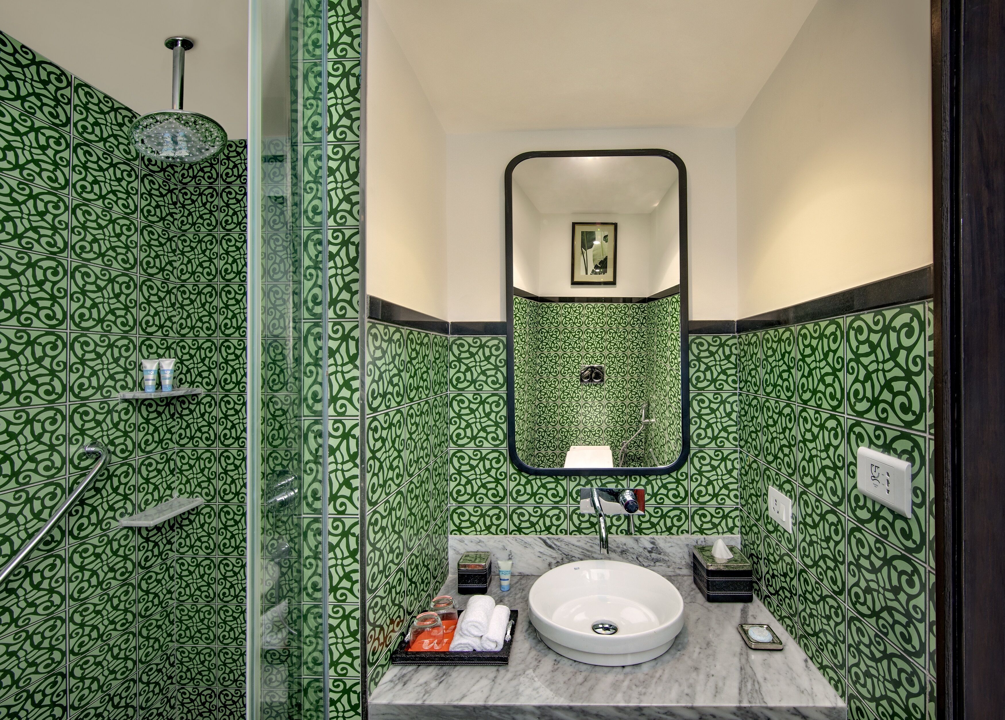 Un baño con baldosas verdes con motivos, un lavabo redondo, un espejo y una zona de ducha.