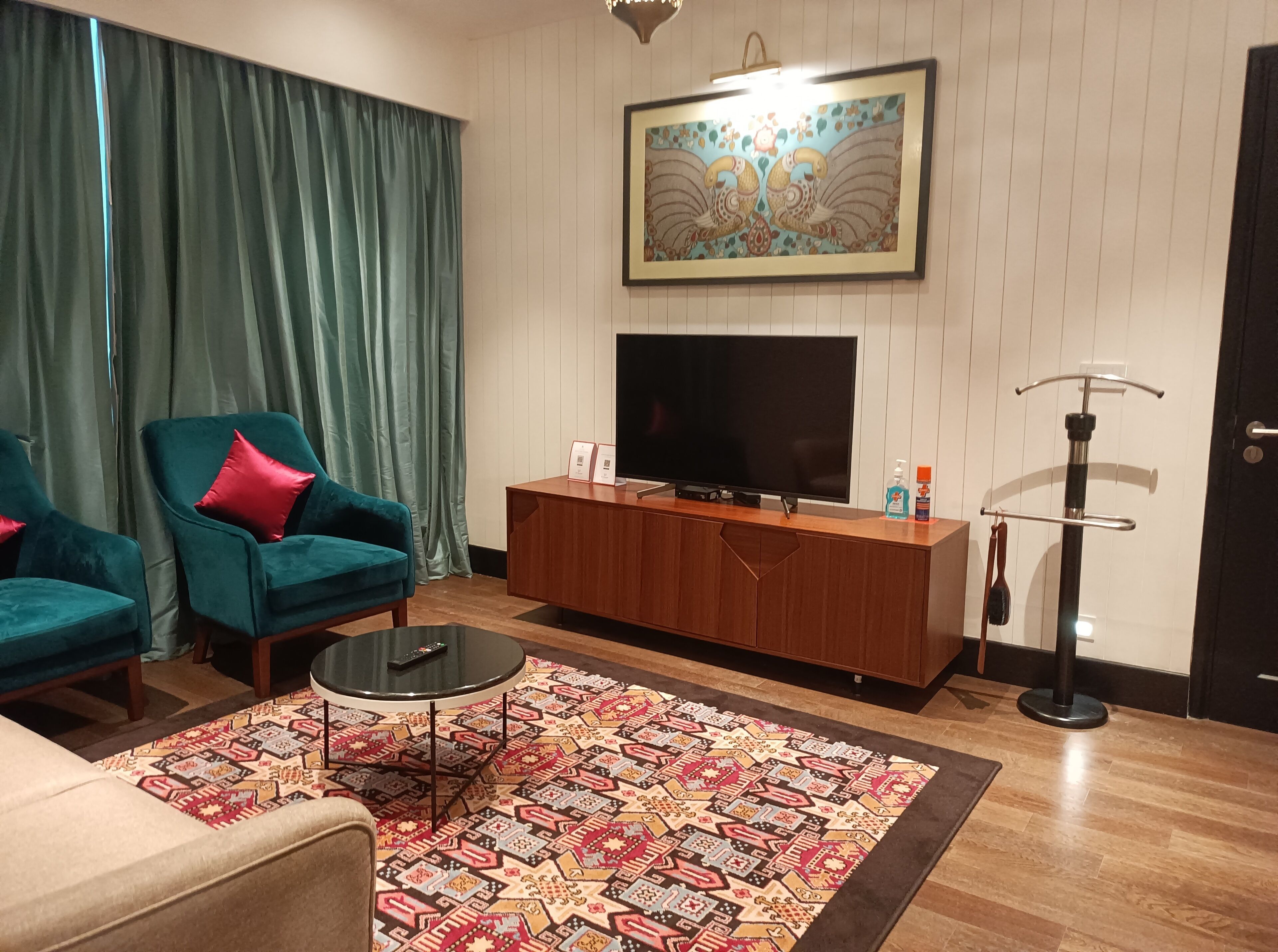A living room with teal armchairs, a wooden TV stand, a patterned rug, and a wall-mounted TV.
