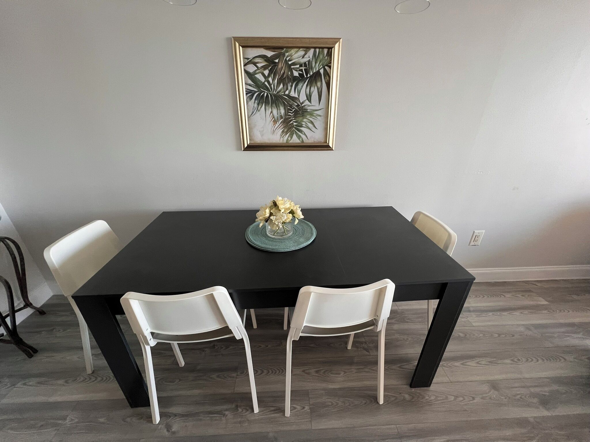 A dining table with a dark surface, surrounded by four white chairs. A framed botanical print hangs on the wall above the table.