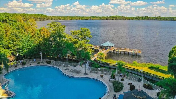 Une piscine dotée d’une terrasse en bois et de chaises longues, entourée d’une végétation luxuriante et bordée d’un quai s’avançant dans un lac.