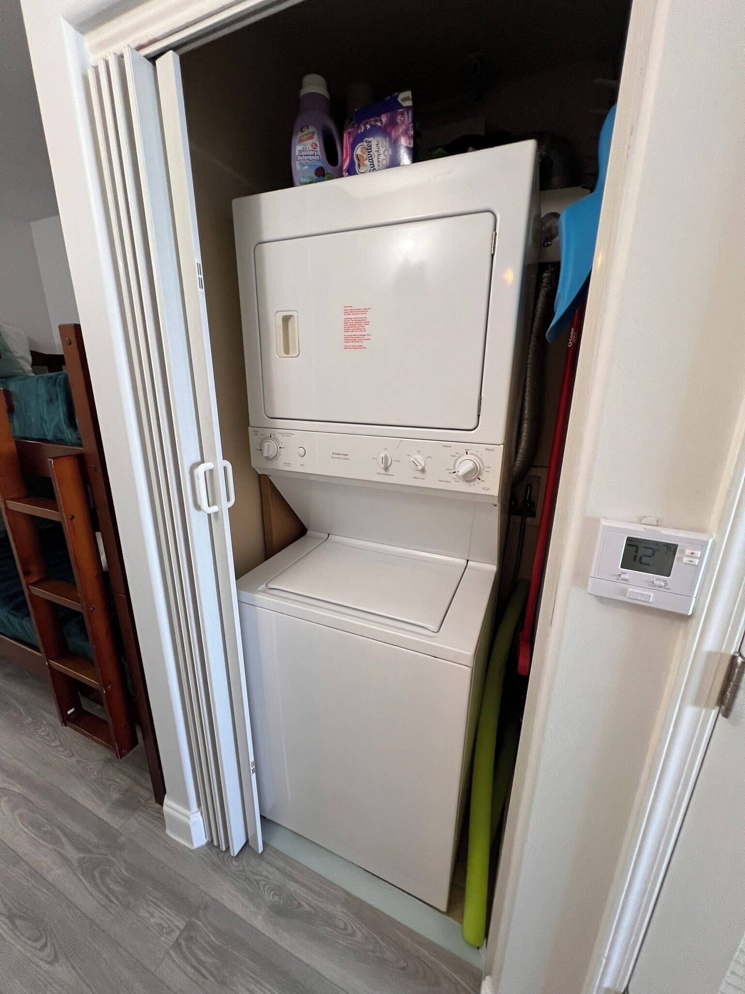Uma lavanderia compacta com máquina de lavar e secar frontal, uma pequena prateleira com produtos de limpeza e um termostato digital na parede.