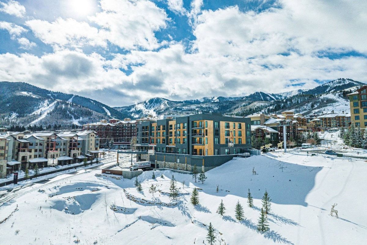 A modern hotel complex with multiple buildings, a ski slope, and snow-covered mountains in the background.