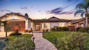 Una casa residencial con un sendero de ladrillo que conduce a la entrada, rodeada de jardines cuidados y una palmera.