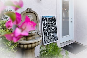 A stone fountain with water spouting from it, a welcome sign for a beach house, and a door with a window and a vent.