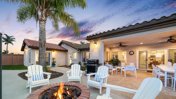 Un patio con un hogar para fogata, sillas Adirondack blancas y un comedor techado.