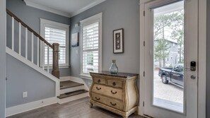 Un escalier avec des rampes blanches et une main courante en bois, menant à une pièce dont un mur est peint en bleu clair, où se trouvent une commode en bois et une porte donnant sur la rue.