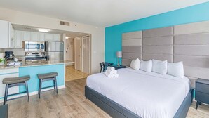 A modern hotel room with a kitchenette, a large bed with a tufted headboard, and a small seating area with stools. - Palace 1501 (Myrtle Beach)