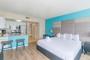 Una habitación de hotel moderna con kitchenette, una cama grande con cabecera capitonada y un pequeño rincón de descanso con taburetes.