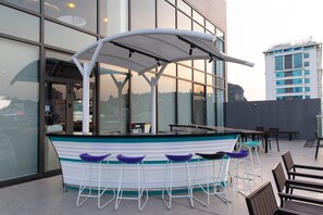 A bar area with a boat-shaped counter and stools, a table with chairs, and a view of a building with large windows. - Tru by Hilton Ha Long Hon Gai Centre (Ha Long)