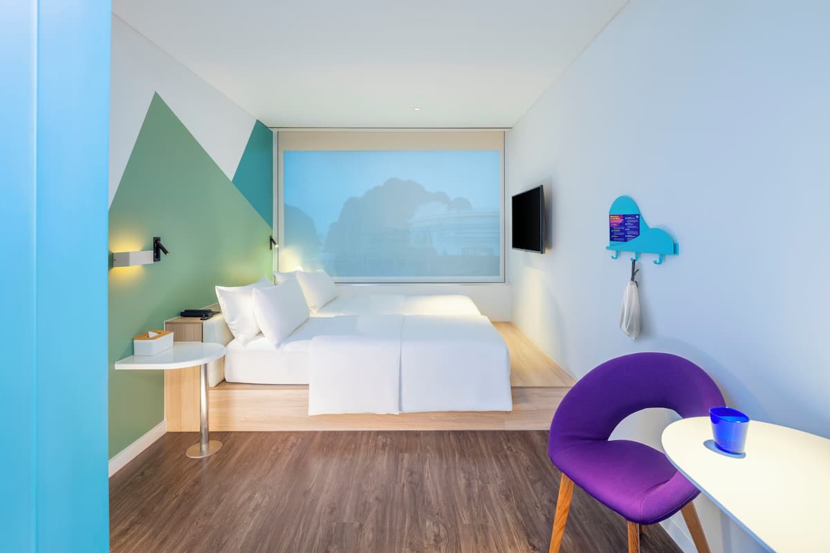 Ein modernes Hotelzimmer mit einem Bett, einem kleinen Tisch, einem violetten Sessel, einem Fernseher und einer Projektionsleinwand an der Wand.