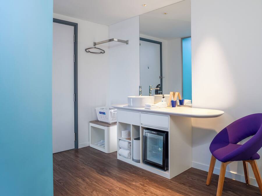 Un baño moderno con un tocador blanco, refrigerador empotrado, una silla morada y un espejo grande.