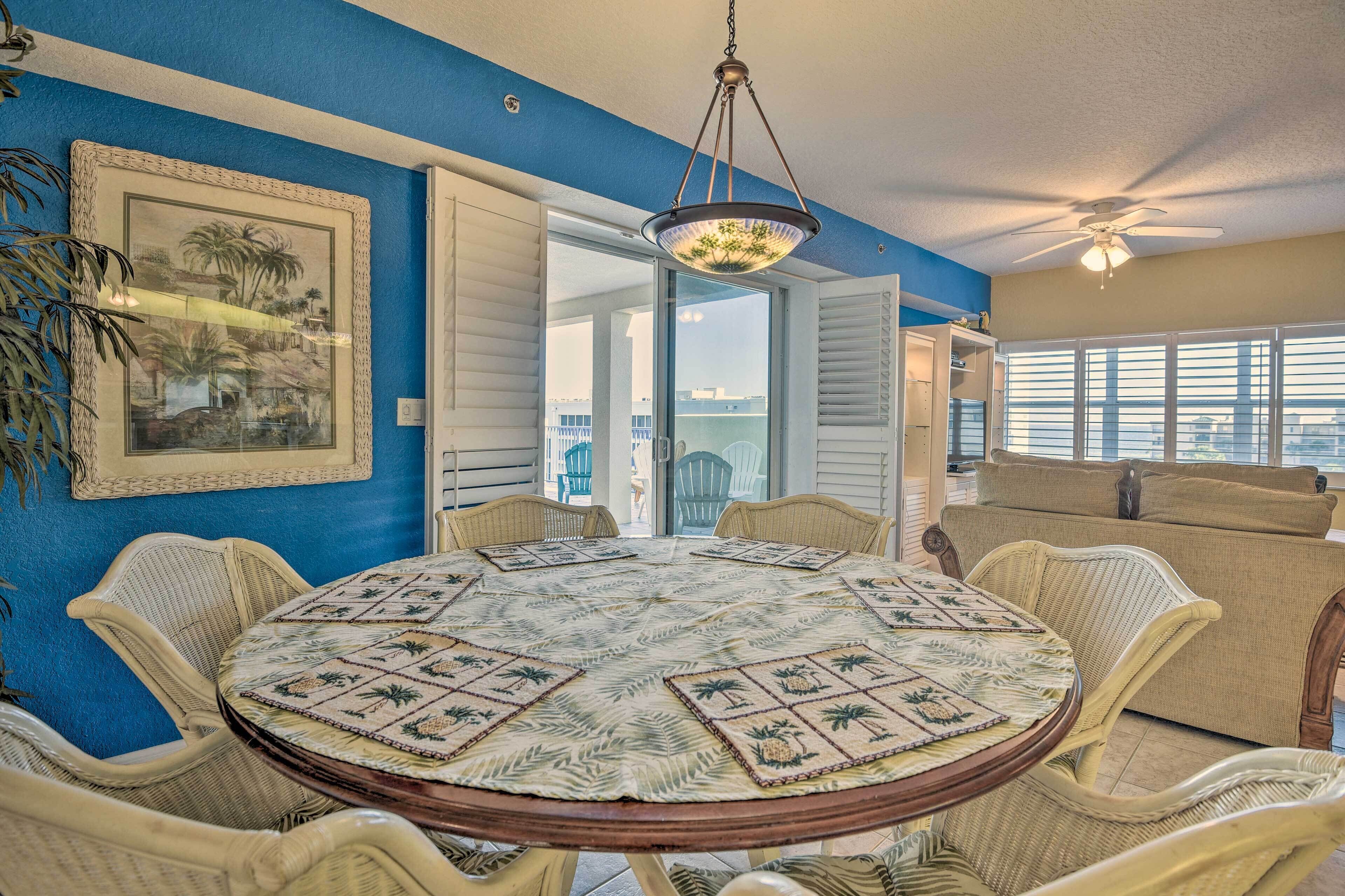 Une table de salle à manger ronde, recouverte d’une nappe aux motifs tropicaux, entourée de chaises en osier, dans une pièce aux murs bleus et dotée d’un ventilateur de plafond.