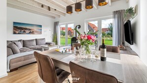 A modern living room with a sofa, dining table, and chairs. There is a painting on the wall, a plant, and a TV mounted on the wall. - Vacation apartment Nordseebrise in the Nordseeresort - terrace, fireplace, infrared cabin, S (Büsum)