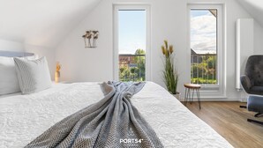 A bedroom with a large bed, two windows, a chair, and a small table. - Vacation apartment Nordseebrise in the Nordseeresort - terrace, fireplace, infrared cabin, S (Büsum)