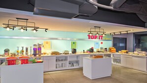 Un área de desayuno moderna con un letrero 'TOP IT', diversas estaciones de comida y un interior limpio y bien iluminado.