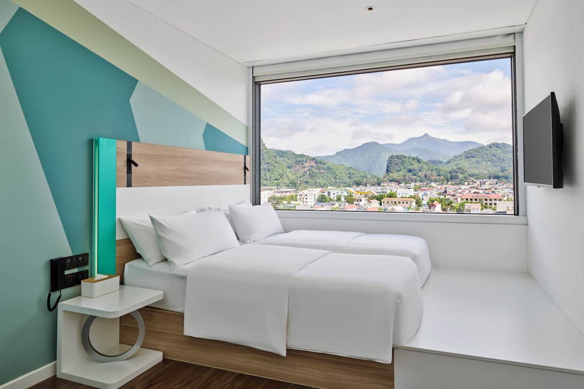 A modern bedroom with a bed, bedside table, and a large window offering a cityscape view.