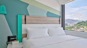 A modern hotel room with a large bed, a bedside table with a tissue box, a wall-mounted phone, and a view of a city and mountains through a large window.