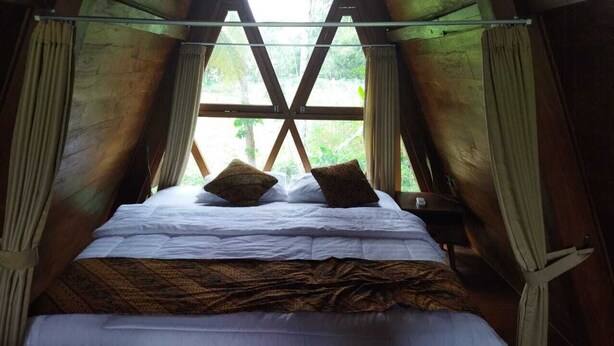 A bed with white linens and two decorative pillows, set against a wooden ceiling with a window above. - Borobudur Balkondes Saka Pitu (Yogyakarta)