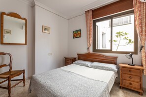 A bedroom with a bed, a wooden chair, a nightstand, a mirror, and a window with curtains. - Apartment "3 Bedrooms 3 Terraces Maquinilla" with Wi-Fi, A/C & Terraces (Nerja)