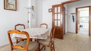 A dining area with a round table, wooden chairs, a lace tablecloth, a framed picture on the wall, and a door leading to a kitchen.