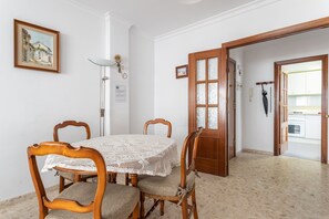 Un coin repas avec une table ronde, des chaises en bois, une nappe en dentelle, un tableau encadré au mur et une porte donnant sur la cuisine.