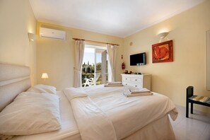 A bedroom with a bed, a TV, a painting, and a window with curtains. - Holiday Home "Primavera" with Sea View, Pool, Wi-Fi, A/C, Terraces & Garden (Carvoeiro)