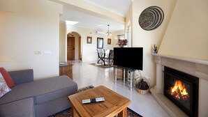 A living room with a sofa, a fireplace, a TV, and a dining area in the background. - Holiday Home "Primavera" with Sea View, Pool, Wi-Fi, A/C, Terraces & Garden (Carvoeiro)