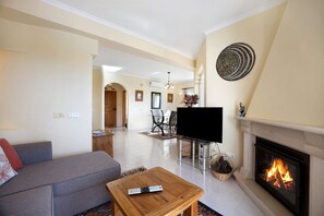 A living room with a sofa, a fireplace, a TV, and a dining area in the background. - Holiday Home "Primavera" with Sea View, Pool, Wi-Fi, A/C, Terraces & Garden (Carvoeiro)