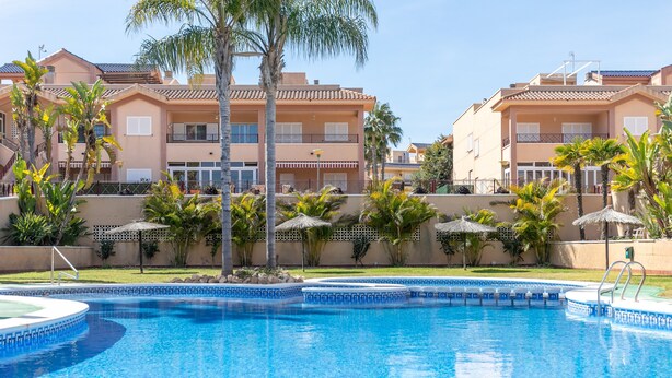 A swimming pool with a clear blue water, surrounded by palm trees and a building with balconies. - Holiday Apartment with Sea View, Shared Pool & Jacuzzi, Terraces, Garden & Wi-Fi (Gran Alacant)