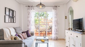 A living room with a sofa, a coffee table with fruit, a TV on a cabinet, and a balcony with a table and chairs. - Holiday Apartment with Sea View, Shared Pool & Jacuzzi, Terraces, Garden & Wi-Fi (Gran Alacant)