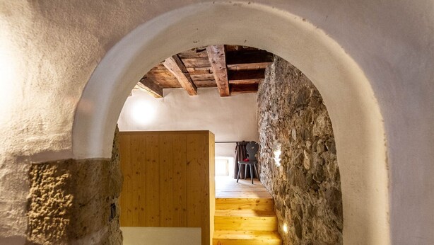 Una escalera de paredes de piedra que lleva a una puerta de madera, con un interior rústico y vistas a una habitación con cama y silla.