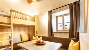 Una habitación con literas, una mesa de comedor con frutero, una ventana con cortinas y vistas a flores afuera.