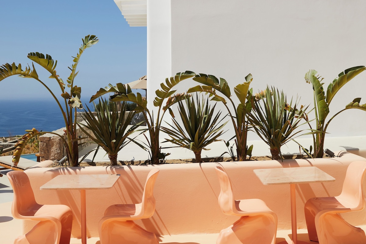 A white building with a door and windows, surrounded by plants and a table with chairs.